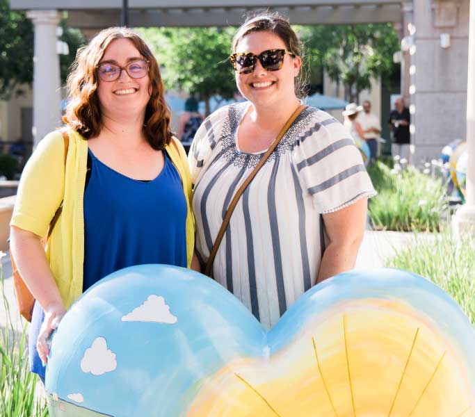 two ladies smiling at a the camera while standing in front of a painted heart sculpture