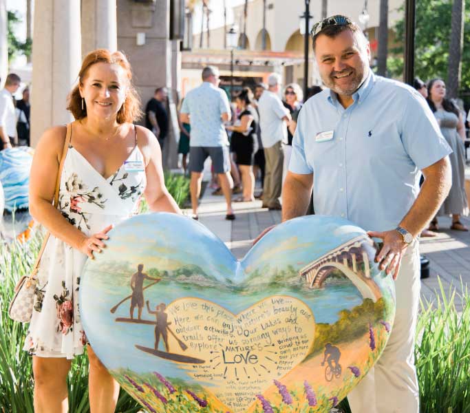 A couple standing in front of painted heart sculpture while smiling at the camera