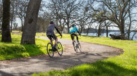 two people riding on a paved path with Folsom lake in front of them.