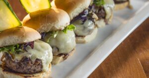 A plate of burger sliders with a pickle standing on top of bun.