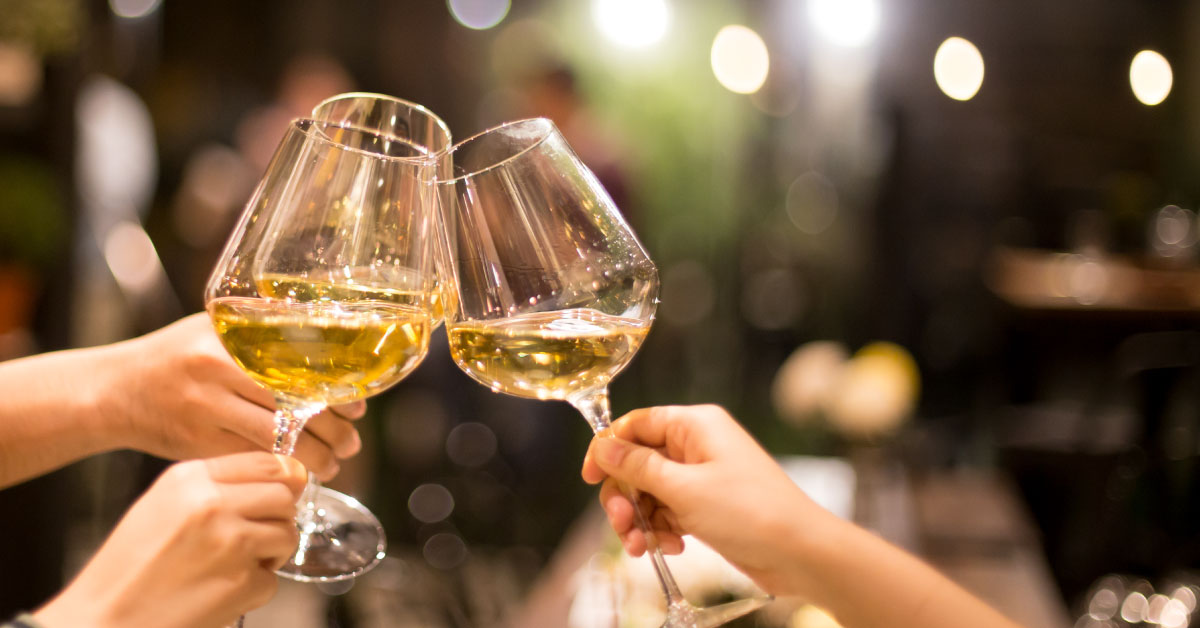 Three hands clinking glasses of white wine in a celebratory toast, with a blurred background of warm lights and an inviting indoor setting.